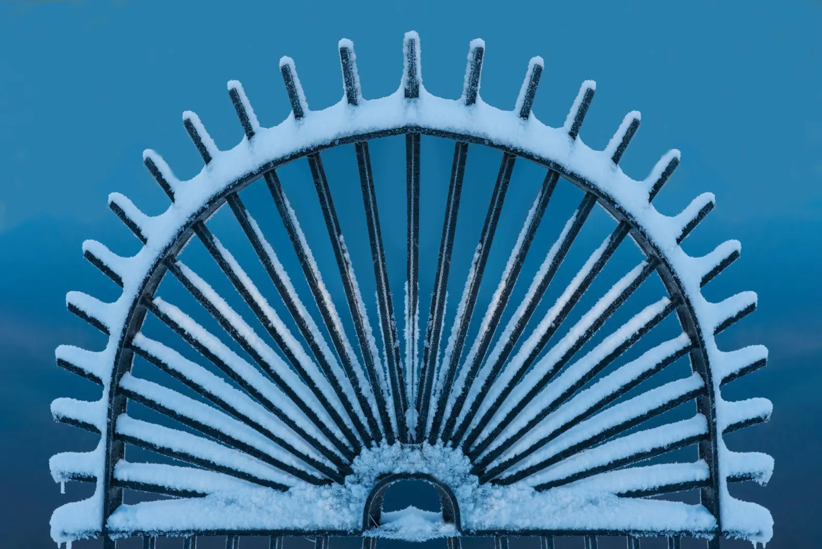 Snow and frost packed onto the arched top rail of an ornamental iron gate against a winter sky