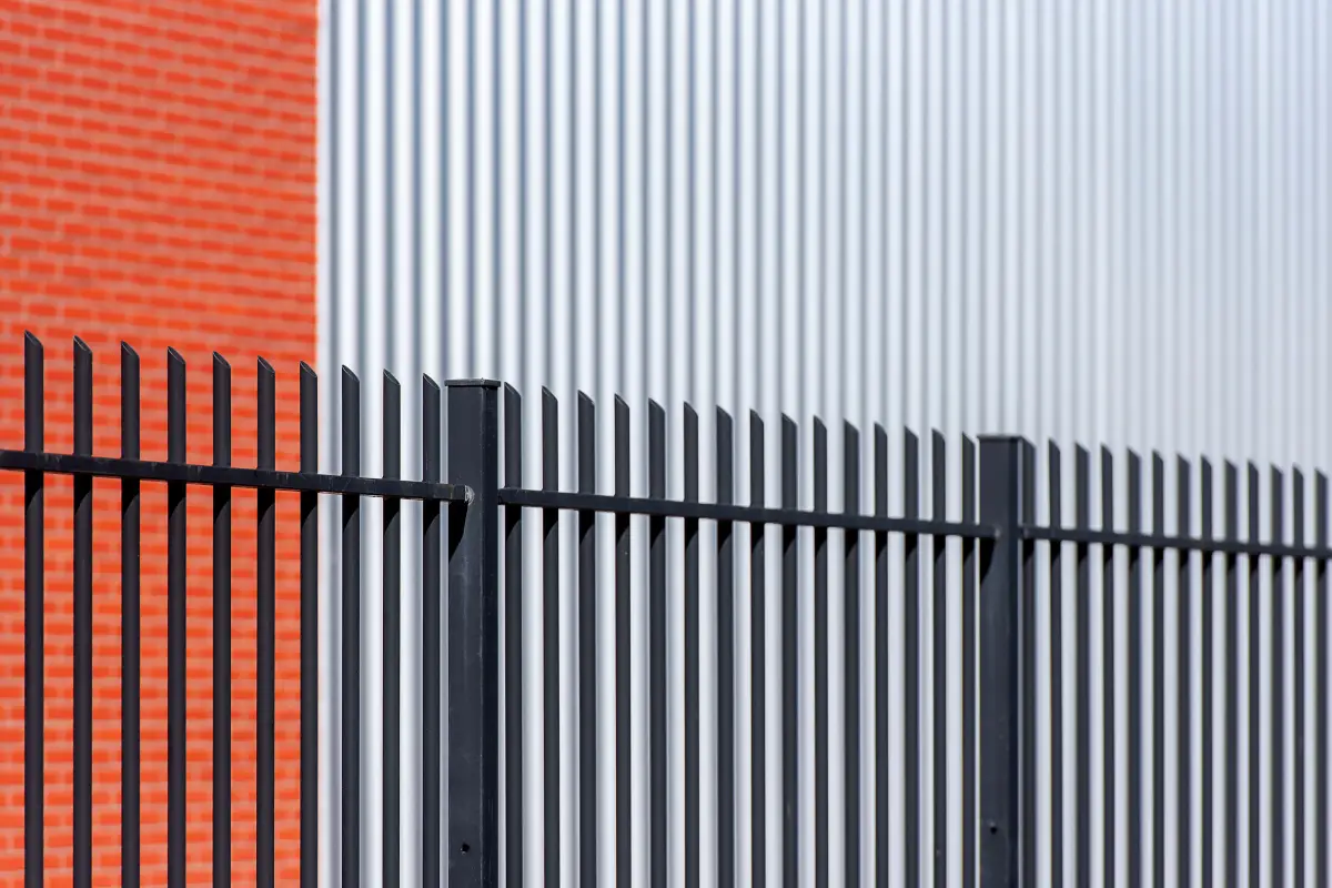 A black steel palisade fence running along a commercial building's brick and metal siding