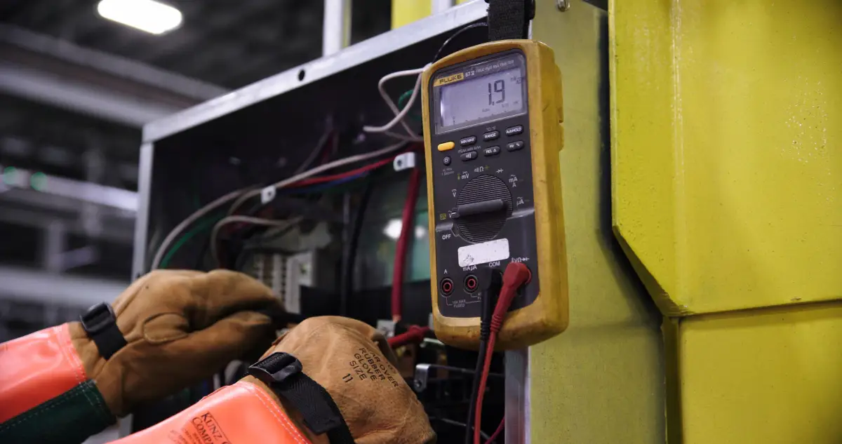 A multimeter reading voltage inside an opened control panel, gloved hands on the probes